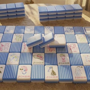 May include: A set of rectangular mahjong tiles featuring a blue and white striped pattern on the back. The front faces display various illustrations including flowers, ribbons, hats, keys, and champagne glasses, with several tiles labeled JOKER.