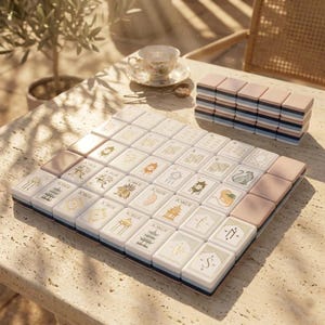 May include: A set of rectangular mahjong tiles with white faces featuring gold and black illustrations of pagodas, animals, and the word Joker. The tiles have pastel pink, blue, and white layered sides, arranged in a square grid on a stone table.