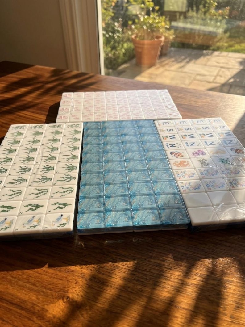May include: A set of rectangular mahjong tiles arranged in three distinct groups. One group features green plant illustrations, another displays a blue swirled pattern, and the third shows various colorful icons and the letters E, S, W, and N.