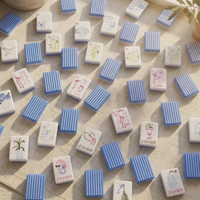 May include: A collection of rectangular mahjong tiles featuring white faces with various illustrations, numbers, and the word JOKER, alongside blue tiles with white vertical stripes. The tiles are scattered across a light-colored fabric surface.