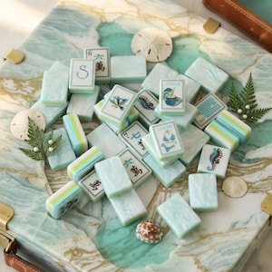 May include: A selection of light blue and white Mahjong tiles featuring diverse symbols and illustrations, such as a seahorse, bikini, and lotus flower. Some tiles display Chinese characters. The tiles are arranged on a marbled surface with decorative elements.