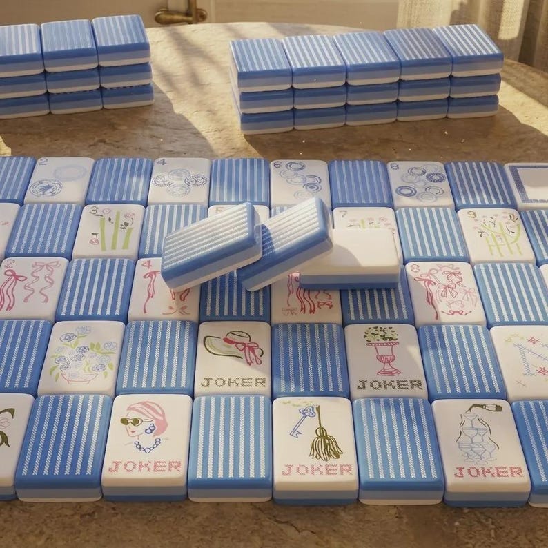 May include: A set of rectangular mahjong tiles featuring a blue and white striped pattern on the back. The front faces display various illustrations including flowers, ribbons, hats, keys, and champagne glasses, with several tiles labeled JOKER.