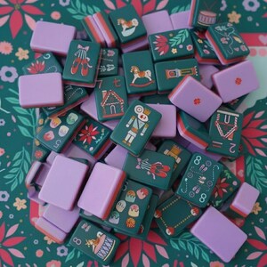 May include: A collection of rectangular mahjong tiles featuring a dark green base with festive illustrations including nutcrackers, ballet slippers, gingerbread houses, and rocking horses, interspersed with solid light purple tiles.