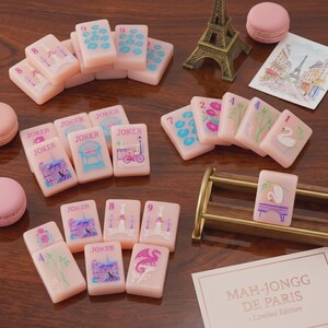 K&ouml;nnte beinhalten: Ein Set aus blassrosa Mahjong-Spielsteinen mit bunten Illustrationen von Pariser Wahrzeichen, Schw&auml;nen und Drachen, erg&auml;nzt durch ein Miniatur-Eiffelturm-Modell, rosa Macarons, eine Postkarte und eine Karte mit der Aufschrift Mah-Jongg De Paris Limited Edition.