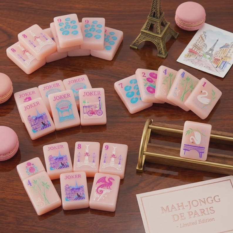 K&ouml;nnte beinhalten: Ein Set aus blassrosa Mahjong-Spielsteinen mit bunten Illustrationen von Pariser Wahrzeichen, Schw&auml;nen und Drachen, erg&auml;nzt durch ein Miniatur-Eiffelturm-Modell, rosa Macarons, eine Postkarte und eine Karte mit der Aufschrift Mah-Jongg De Paris Limited Edition.