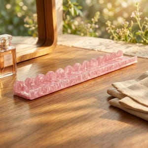Pode incluir: Um expositor retangular longo de acr&iacute;lico rosa com uma s&eacute;rie de entalhes arredondados e recortados ao longo da borda superior. O material tem uma apar&ecirc;ncia marmorizada e transl&uacute;cida com um acabamento brilhante.