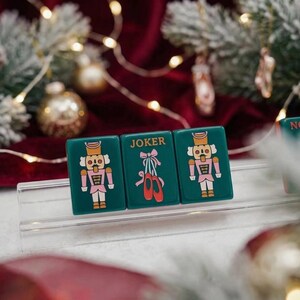 May include: Three rectangular dark green tiles displayed on a clear stand. The outer tiles feature a nutcracker illustration in pink and white. The center tile displays the word JOKER above an illustration of pink ballet slippers with red ribbons.