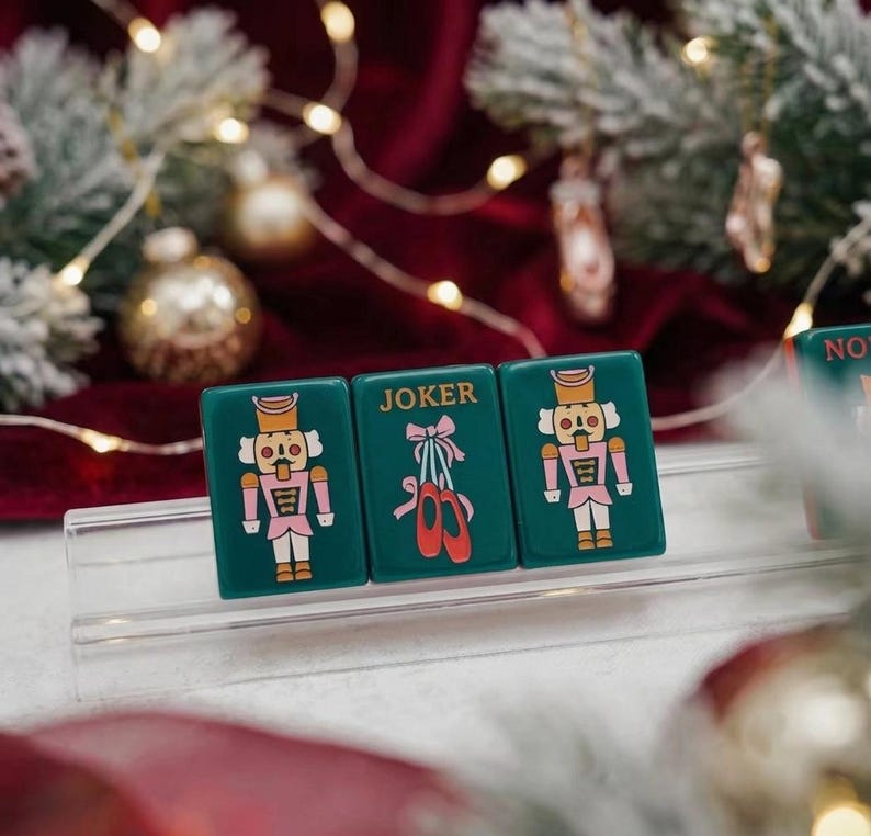 May include: Three rectangular dark green tiles displayed on a clear stand. The outer tiles feature a nutcracker illustration in pink and white. The center tile displays the word JOKER above an illustration of pink ballet slippers with red ribbons.