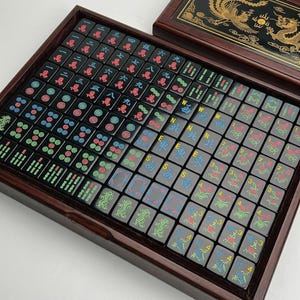 May include: A set of black Mahjong tiles arranged in a dark wooden box. The tiles feature colorful engraved Chinese characters, numbers, and symbols in red, blue, and green. The box lid displays a gold dragon and phoenix design on a black background.