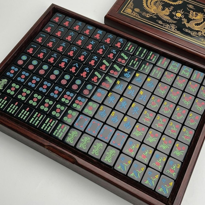 May include: A set of black Mahjong tiles arranged in a dark wooden box. The tiles feature colorful engraved Chinese characters, numbers, and symbols in red, blue, and green. The box lid displays a gold dragon and phoenix design on a black background.