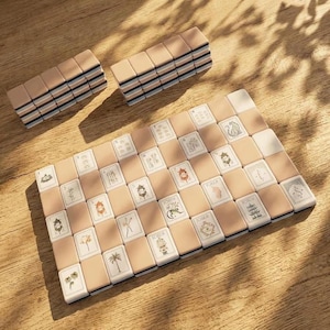 May include: A mahjong set featuring rectangular tiles with beige backs and white faces. The tiles display various illustrations including pagodas, palm trees, flowers, and the word Joker. Some tiles are arranged in a grid, while others are stacked.
