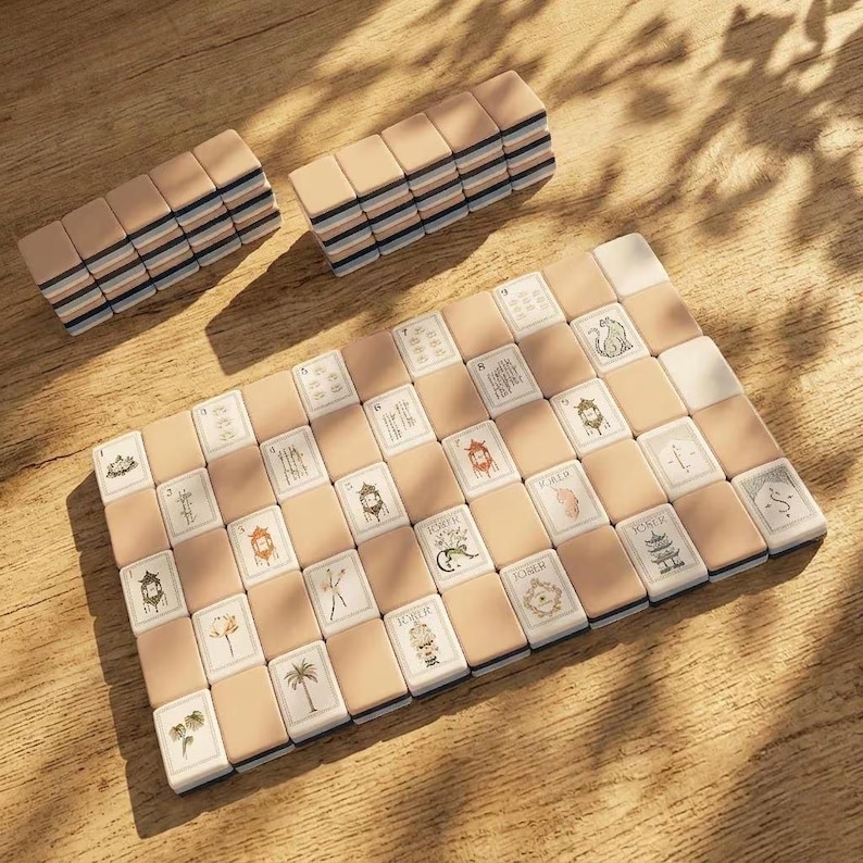 May include: A mahjong set featuring rectangular tiles with beige backs and white faces. The tiles display various illustrations including pagodas, palm trees, flowers, and the word Joker. Some tiles are arranged in a grid, while others are stacked.