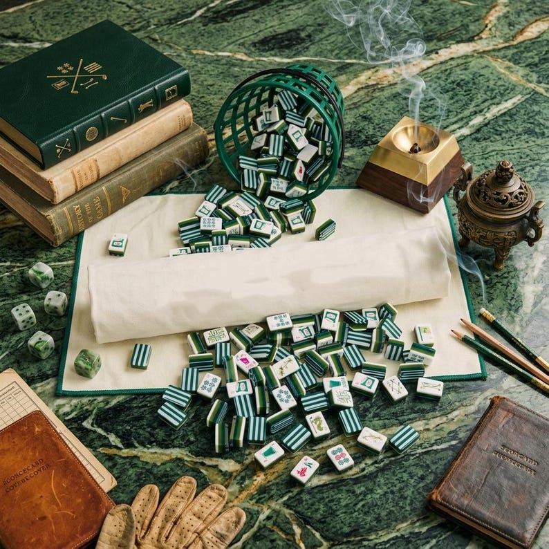 May include: A Mahjong set with green and white tiles spilling from a green basket onto a cream-colored mat. The scene includes a stack of three books, a brass incense burner with smoke, a small bronze censer, dice, and a brown leather scorebook.