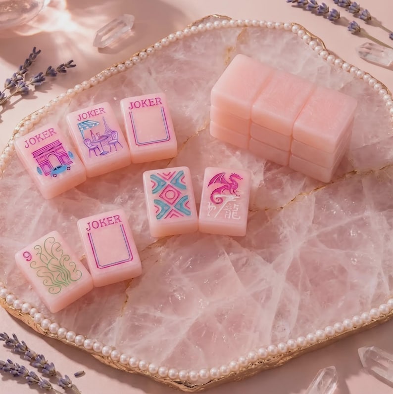 K&ouml;nnte beinhalten: Ein Set aus blassrosa Mahjong-Spielsteinen mit verschiedenen eingravierten Motiven, darunter das Wort JOKER, ein Drache, ein Blumenmuster und eine Pariser Szene. Die Steine liegen auf einer gro&szlig;en, unregelm&auml;&szlig;igen Platte aus durchscheinendem Rosenquarz mit Perlenrand.