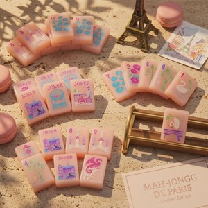 K&ouml;nnte beinhalten: Ein Set hellrosa Mahjong-Spielsteine mit bunten Illustrationen von Pariser Wahrzeichen, Fahrr&auml;dern, Schw&auml;nen und Drachen, dazu ein Miniatur-Eiffelturm, rosa Macarons und eine Karte mit der Aufschrift Mah-Jongg de Paris Limited Edition.