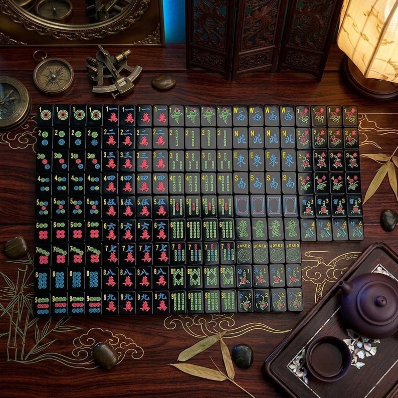 May include: A complete set of black mahjong tiles arranged in a grid, featuring colorful engraved Chinese characters, numbers, and symbols in red, blue, green, and yellow, including directional markers and joker tiles on a dark wooden surface.