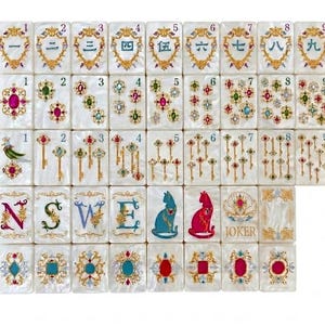 K&ouml;nnte beinhalten: Ein Set aus wei&szlig;en, perlmuttfarbenen Mahjong-Spielsteinen mit goldenen Filigranrahmen. Die Steine zeigen chinesische Ziffern von eins bis neun, bunte Edelsteincluster, goldene Schl&uuml;ssel, die Buchstaben N, S, W, E, Katzensilhouetten und einen Joker.