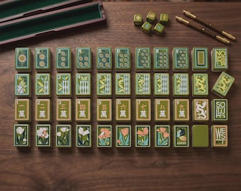May include: A set of rectangular mahjong tiles with green, tan, and white floral, numerical, and dragon designs. The set includes two wooden storage cases, two brown metal sticks, and five small green dice arranged on a dark wooden surface.