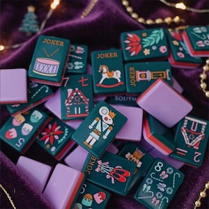 May include: A collection of rectangular mahjong tiles featuring dark green faces with colorful holiday-themed illustrations including a nutcracker, gingerbread house, drum, and ballet slippers, paired with solid light purple backs.