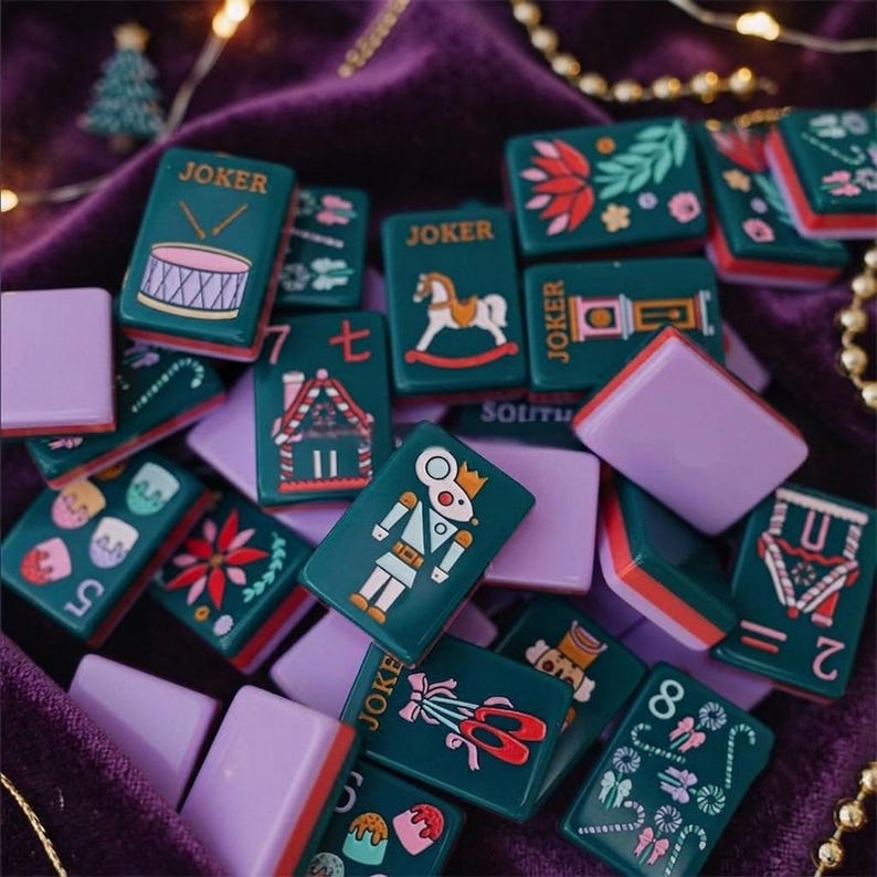 May include: A collection of rectangular mahjong tiles featuring dark green faces with colorful holiday-themed illustrations including a nutcracker, gingerbread house, drum, and ballet slippers, paired with solid light purple backs.