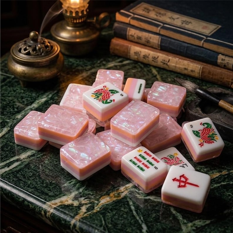 Puede incluir: Una colecci&oacute;n de fichas de mahjong rectangulares con un acabado jaspeado en rosa nacarado y blanco. Varias fichas muestran coloridas ilustraciones grabadas de p&aacute;jaros, flores y caracteres chinos rojos en sus superficies superiores.