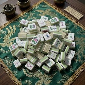 May include: A collection of rectangular mahjong tiles with white faces and light green backs. The tiles feature various colorful illustrations, including floral patterns, Chinese characters, and the letters M, N, and S, arranged in a scattered pile.