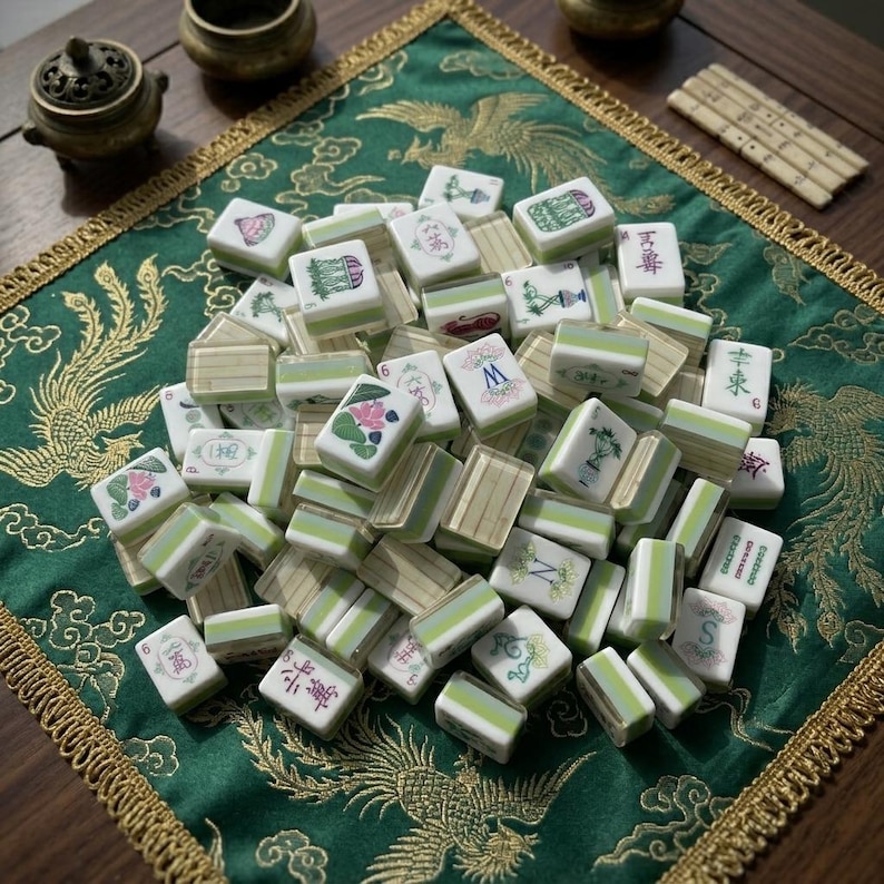 May include: A collection of rectangular mahjong tiles with white faces and light green backs. The tiles feature various colorful illustrations, including floral patterns, Chinese characters, and the letters M, N, and S, arranged in a scattered pile.
