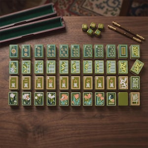 May include: A set of rectangular mahjong tiles with green, tan, and white floral, numerical, and dragon designs. The set includes two wooden storage cases, two brown metal sticks, and five small green dice arranged on a dark wooden surface.