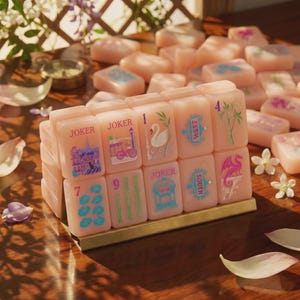 K&ouml;nnte beinhalten: Ein Set aus blassrosa Mahjong-Spielsteinen, die auf einem goldfarbenen Metallst&auml;nder angeordnet sind. Die Steine zeigen bunte Illustrationen wie einen Schwan, einen Drachen, Bambusst&auml;be sowie den Text JOKER, WEST und SOUTH. Weitere Steine liegen verstreut.