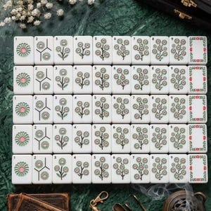 Puede incluir: Un conjunto de fichas de mahjong rectangulares blancas dispuestas en cinco filas, cada una con dise&ntilde;os florales grabados en tonos verdes y rosas, numeradas del uno al nueve, junto a una ficha de borde, sobre una superficie verde oscuro.