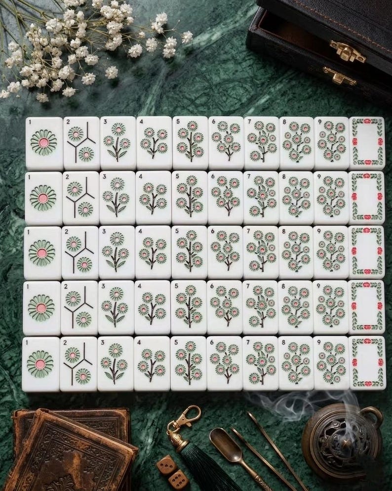 Puede incluir: Un conjunto de fichas de mahjong rectangulares blancas dispuestas en cinco filas, cada una con dise&ntilde;os florales grabados en tonos verdes y rosas, numeradas del uno al nueve, junto a una ficha de borde, sobre una superficie verde oscuro.