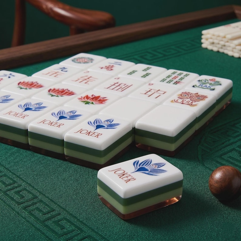 Pode incluir: Um conjunto de pe&ccedil;as retangulares de mahjong com topos brancos e bases verdes em camadas. As pe&ccedil;as apresentam v&aacute;rias ilustra&ccedil;&otilde;es coloridas, incluindo flores de l&oacute;tus azuis, vermelhas, bambu verde e o texto JOKER impresso em vermelho.