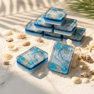 May include: A collection of rectangular resin blocks featuring a marbled pattern of blue, white, and gold glitter. Each block has a layered base of solid blue and white. The blocks are arranged on a sandy surface with various small seashells.