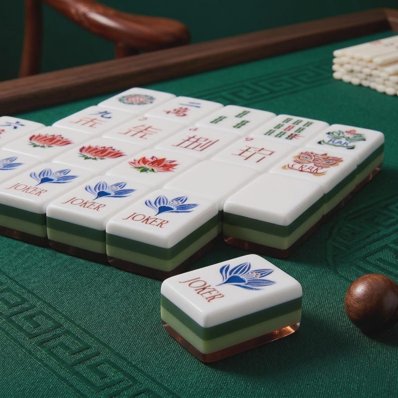 May include: A set of rectangular mahjong tiles with white tops and layered green bases. The tiles feature various colorful illustrations including blue lotus flowers, red lotus flowers, green bamboo, and the text JOKER printed in red with a blue flower icon.