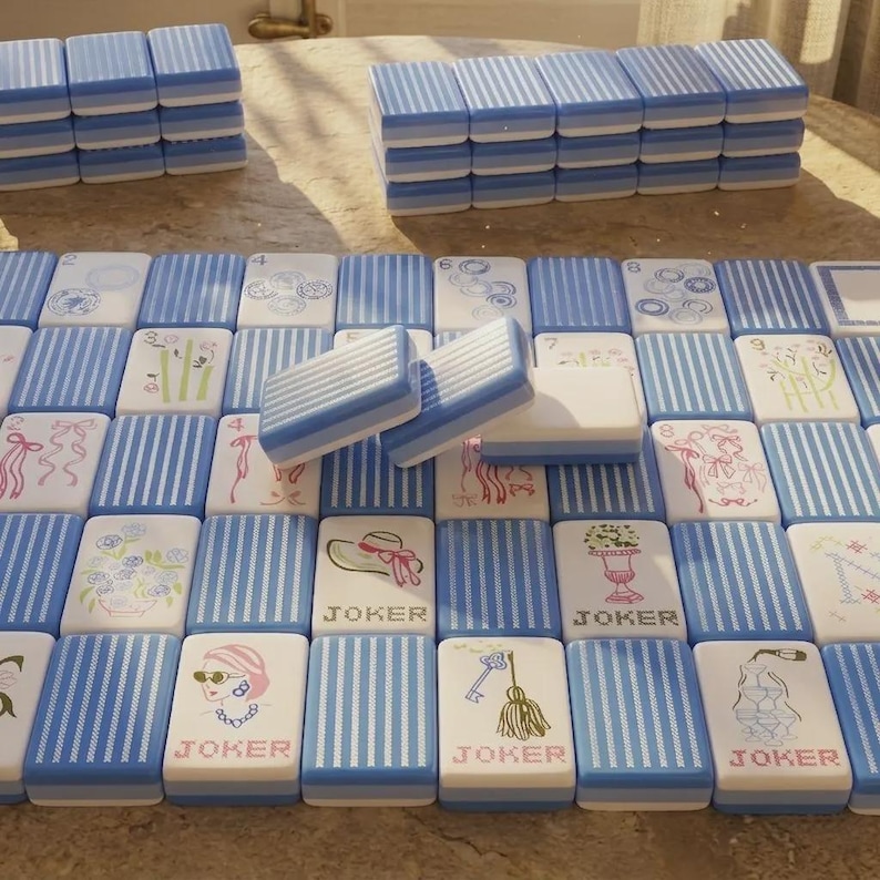May include: A set of rectangular mahjong tiles featuring a blue and white striped pattern on the back. The front faces display various illustrations including flowers, ribbons, hats, keys, and champagne glasses, with several tiles labeled JOKER.