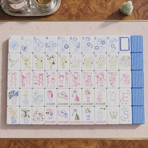 May include: A set of white mahjong tiles arranged in a grid, featuring blue, green, and pink illustrations of flowers, ribbons, birds, hats, and keys. The tiles include numbered suits, joker tiles, and directional markers labeled N, E, W, S.