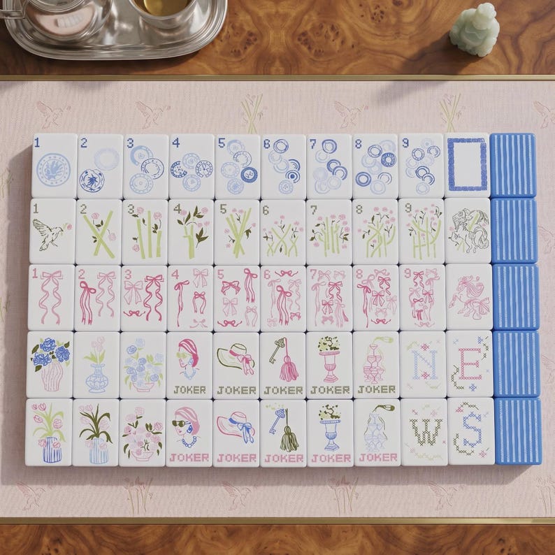 May include: A set of white mahjong tiles arranged in a grid, featuring blue, green, and pink illustrations of flowers, ribbons, birds, hats, and keys. The tiles include numbered suits, joker tiles, and directional markers labeled N, E, W, S.