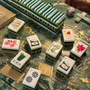 May include: A set of rectangular mahjong tiles featuring white faces with colorful illustrations of flowers, dragons, and joker symbols, alongside green and white striped backs. The tiles are arranged on a green surface with gold scroll patterns.
