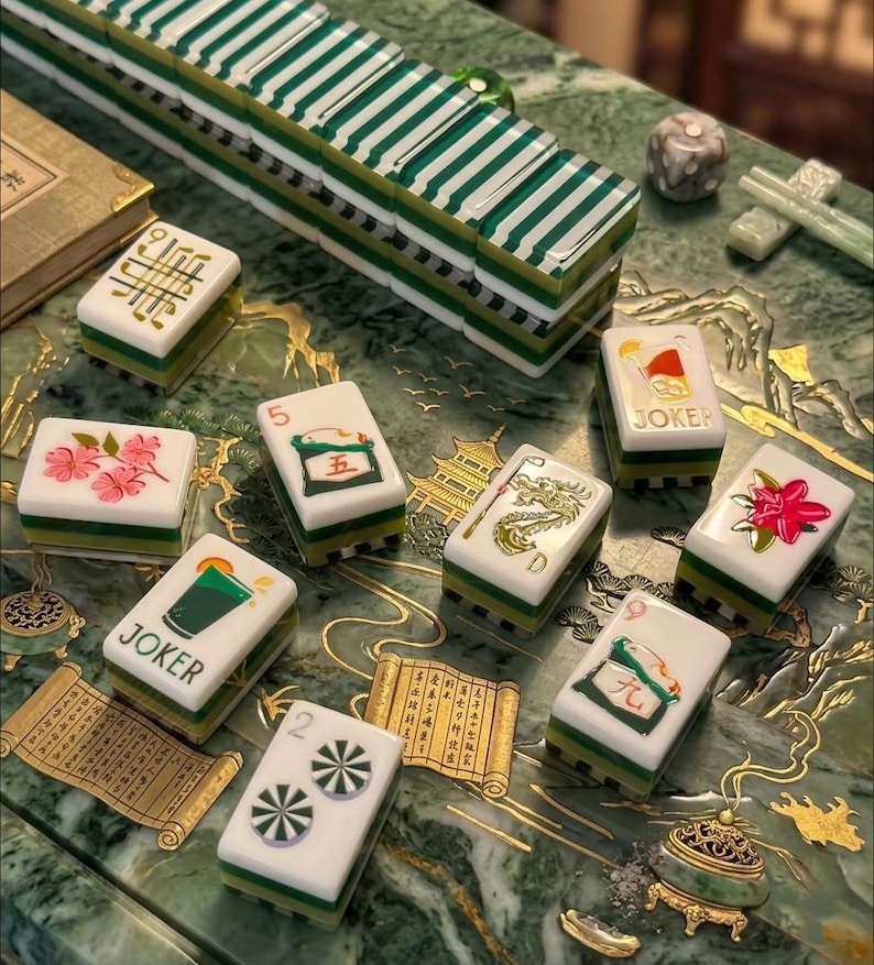 May include: A set of rectangular mahjong tiles featuring white faces with colorful illustrations of flowers, dragons, and joker symbols, alongside green and white striped backs. The tiles are arranged on a green surface with gold scroll patterns.