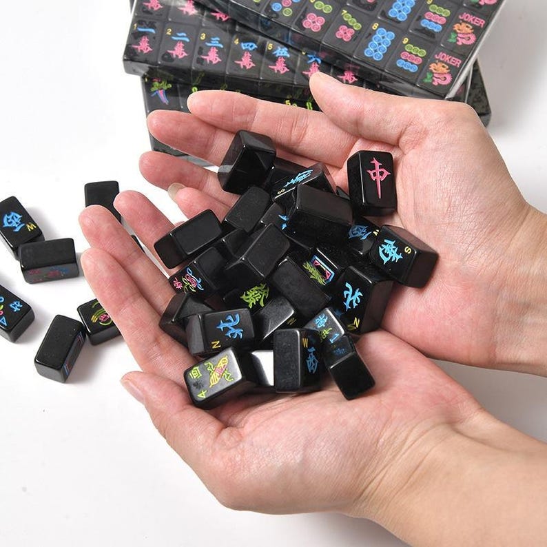 May include: A collection of black mahjong tiles featuring colorful engraved symbols, numbers, and letters. The tiles have a glossy finish with vibrant pink, blue, green, and yellow paint filling the etched characters, including directional markers.