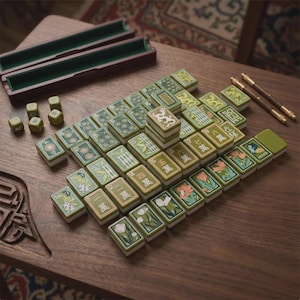 May include: A set of green and white mahjong tiles featuring various floral, bamboo, and numerical designs, arranged on a wooden surface. The set includes a dark wooden storage box, four small green dice, and two thin metal rods with gold accents.