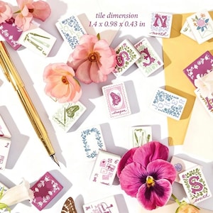 K&ouml;nnte beinhalten: Eine Sammlung rechteckiger Kacheln mit verschiedenen Blumen-, Buchstaben- und Schmetterlingsillustrationen, begleitet von einem goldenen F&uuml;llfederhalter und rosa Bl&uuml;ten. Die Kacheln messen 3,56 x 2,49 x 1,09 cm, wie im Text angegeben.