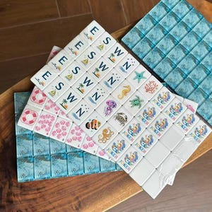 May include: A set of white mahjong tiles featuring various colorful illustrations including sea creatures, palm trees, life preservers, and the word JOKER. The tiles are arranged in rows and columns, with some tiles displaying the letters E, S, W, and N.