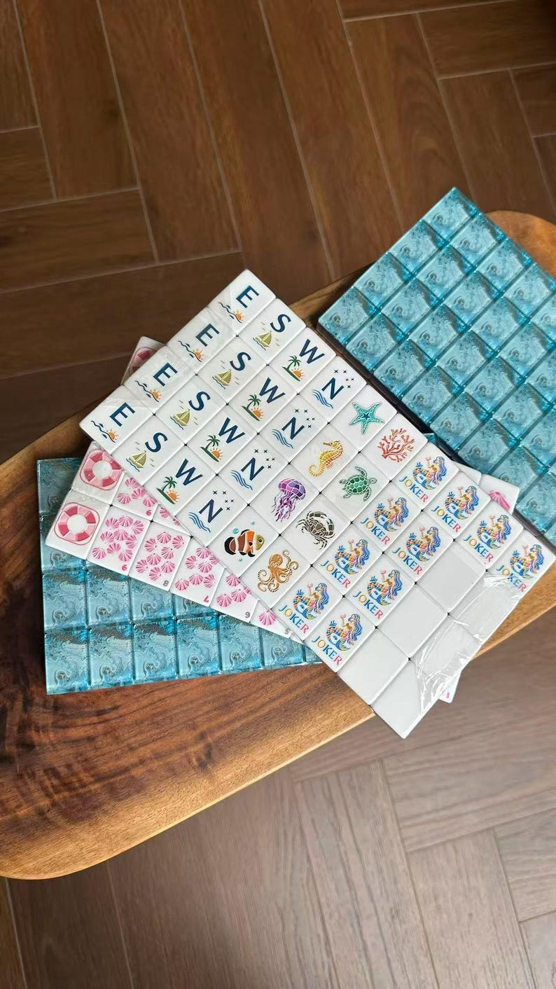 May include: A set of white mahjong tiles featuring various colorful illustrations including sea creatures, palm trees, life preservers, and the word JOKER. The tiles are arranged in rows and columns, with some tiles displaying the letters E, S, W, and N.