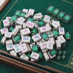 May include: A set of mahjong tiles featuring white faces with pink, green, and blue floral or character designs, paired with translucent emerald green backs. The collection includes several small green dice and four directional tiles.