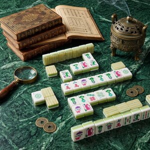 May include: A set of ivory-colored mahjong tiles with green backs and colorful character, bamboo, and joker designs. The tiles are arranged alongside three stacked antique books, a brass magnifying glass, two bronze coins, and a metal incense burner.