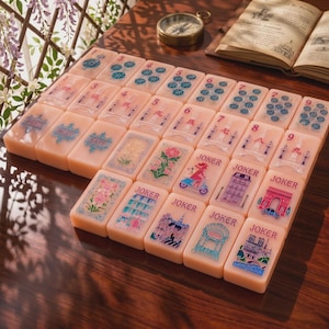 Peut inclure: Un ensemble de tuiles de mah-jong rectangulaires de couleur pêche pâle. Les tuiles présentent divers motifs imprimés, notamment des points numériques bleus, des étiquettes directionnelles comme West et East, des illustrations florales et des Jokers.