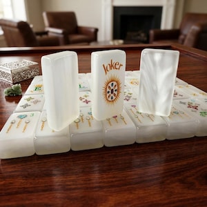 K&ouml;nnte beinhalten: Ein Set aus wei&szlig;en, perlmuttfarbenen Mahjong-Spielsteinen auf einer Holzoberfl&auml;che. Drei Spielsteine stehen aufrecht, wobei der mittlere Stein das Wort Joker in goldener Schrift &uuml;ber einem bunten, sonnenf&ouml;rmigen Edelsteindesign zeigt.