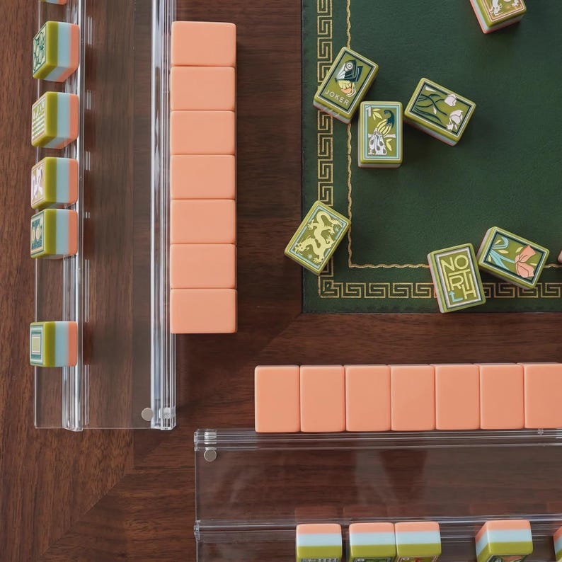 May include: A Mahjong set featuring peach-colored tiles and patterned green tiles with illustrations of dragons, flowers, and the word NORTH. The game includes clear acrylic tile racks and is arranged on a dark wood surface with a green felt mat.