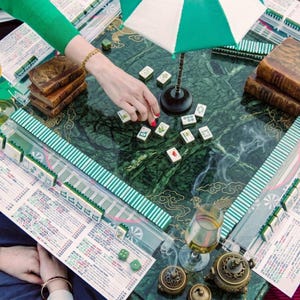 May include: A green marble mahjong table featuring a central green and white striped umbrella, scattered mahjong tiles, and four clear plastic tile racks with printed score sheets. Two antique leather-bound books and a small brass incense burner are present.
